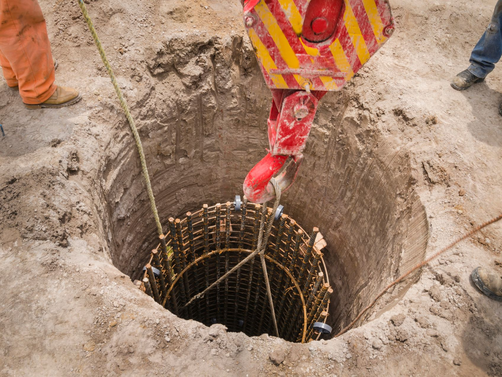 Crane lowering a steel rebar cage into a deep hole in the ground at a construction site.