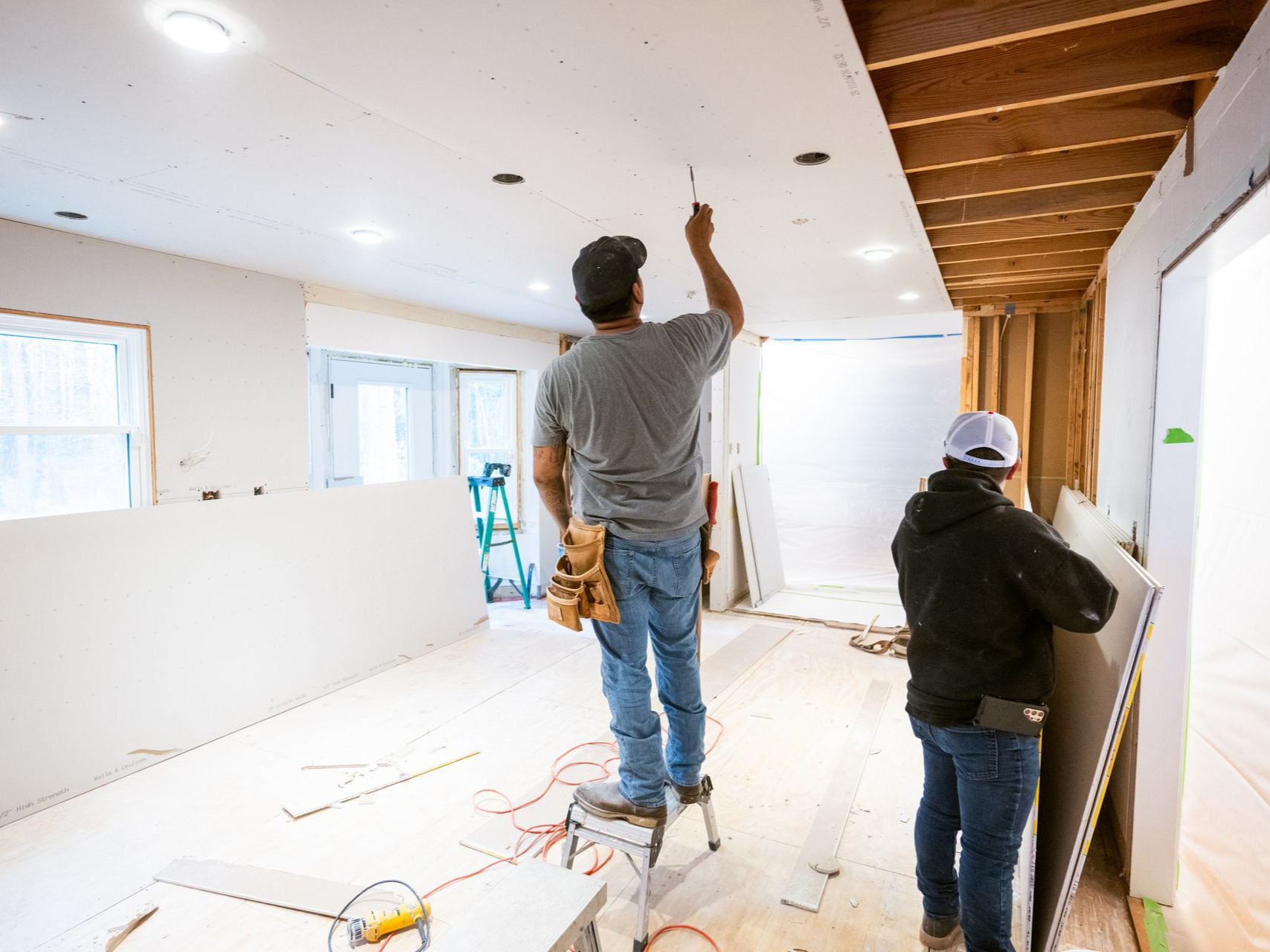 Two workers installing drywall on a room's ceiling. One stands on a stool; the other carries a panel.
