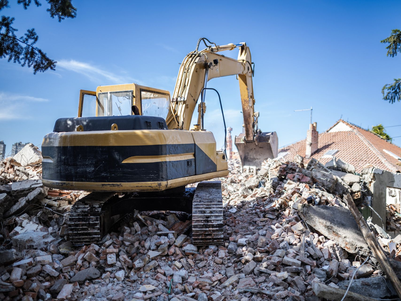 Yellow excavator demolishing a house, debris pile, blue sky.