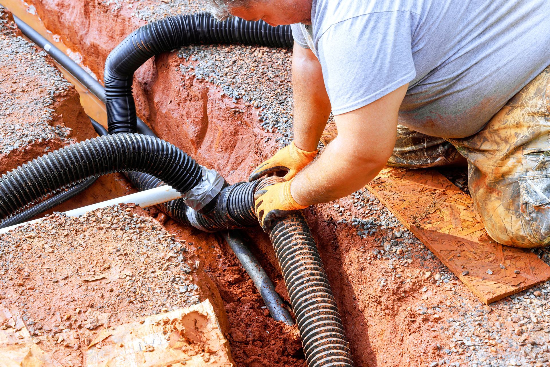 Man connecting black corrugated drainage pipes in a red-soil trench.