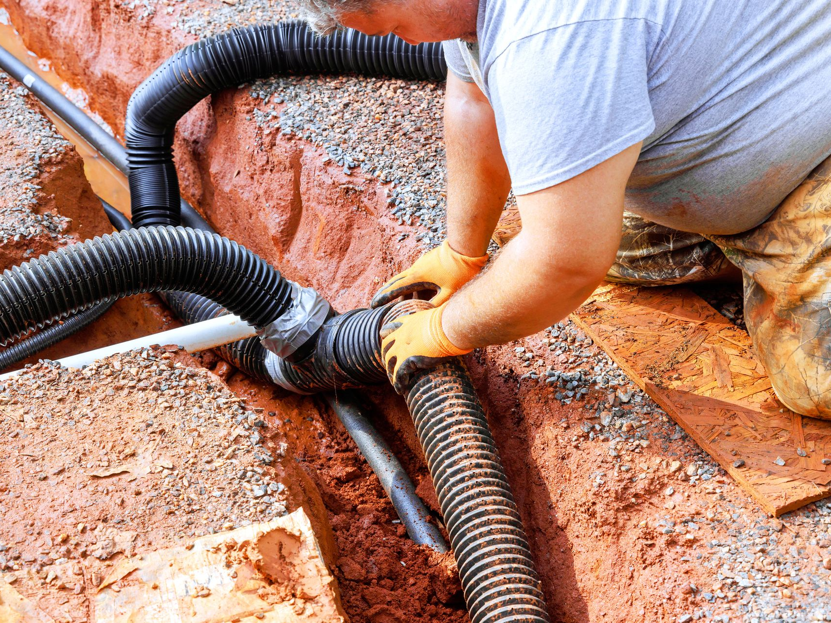 Man connecting corrugated drainage pipes in a trench filled with gravel.