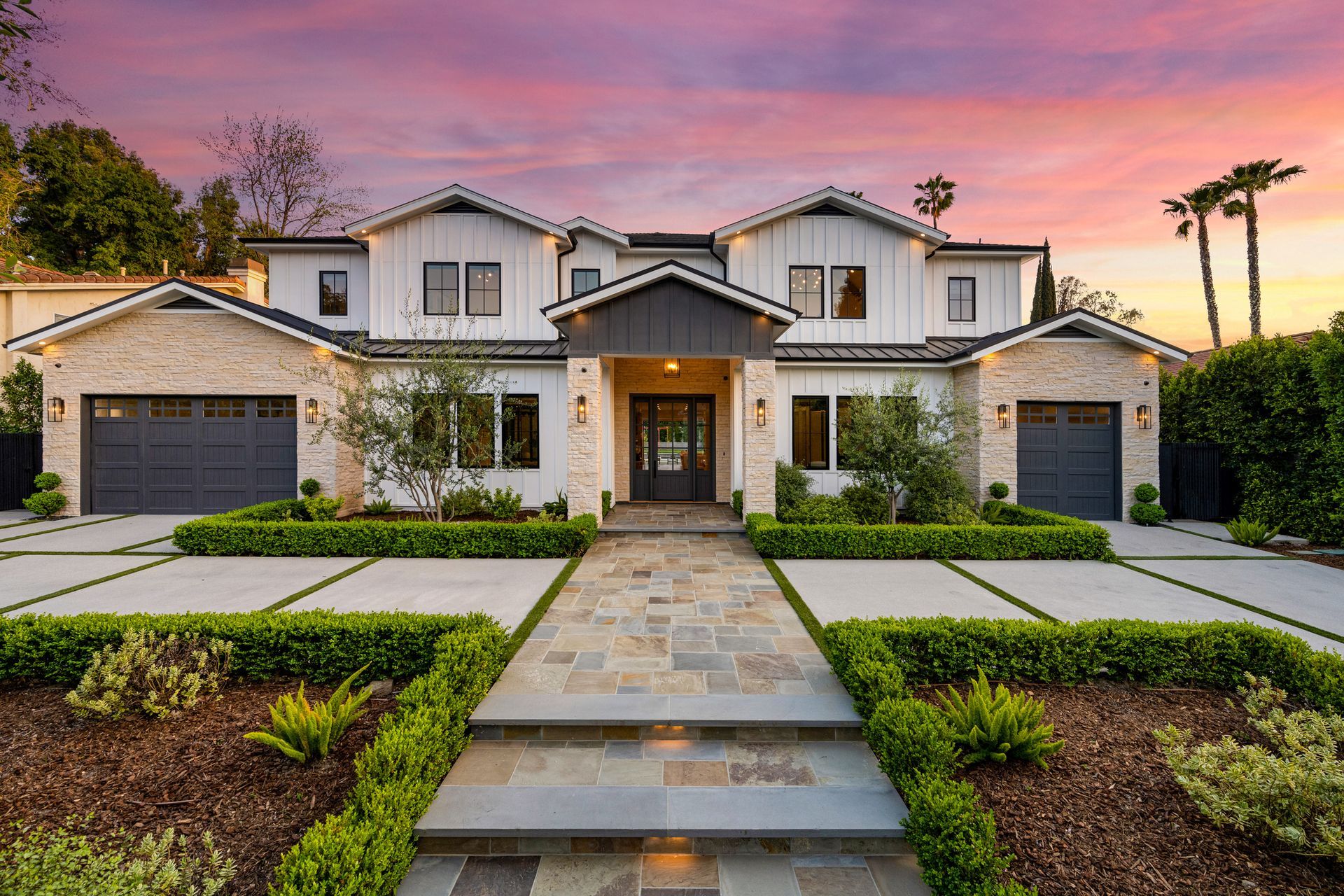 Two-story modern home with stone and white siding, surrounded by landscaping, under a colorful sunset sky.
