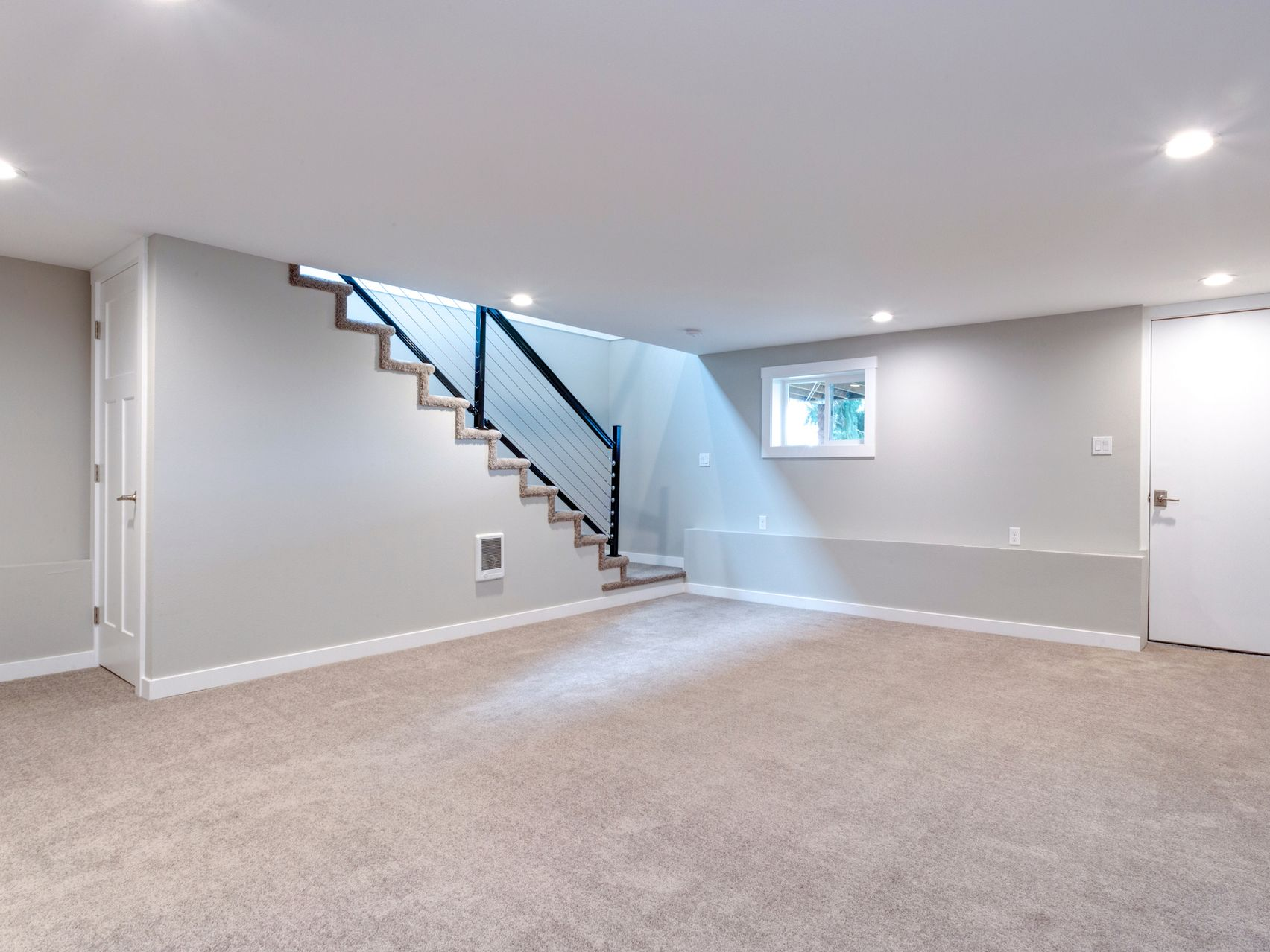 Empty basement with gray walls, carpet, and staircase with black railing.