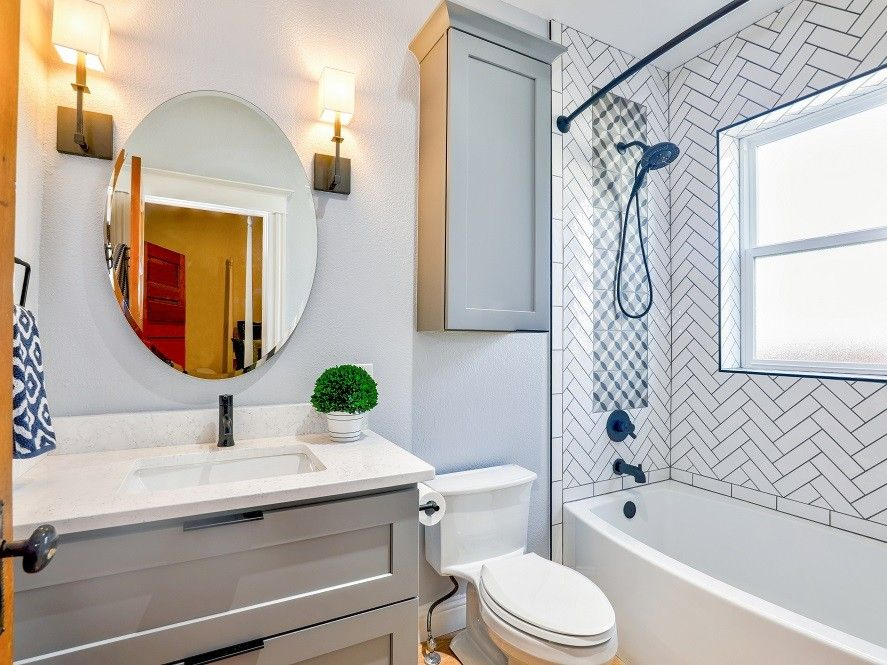 Bathroom with gray vanity, round mirror, white tile, bathtub, and gray storage cabinet.