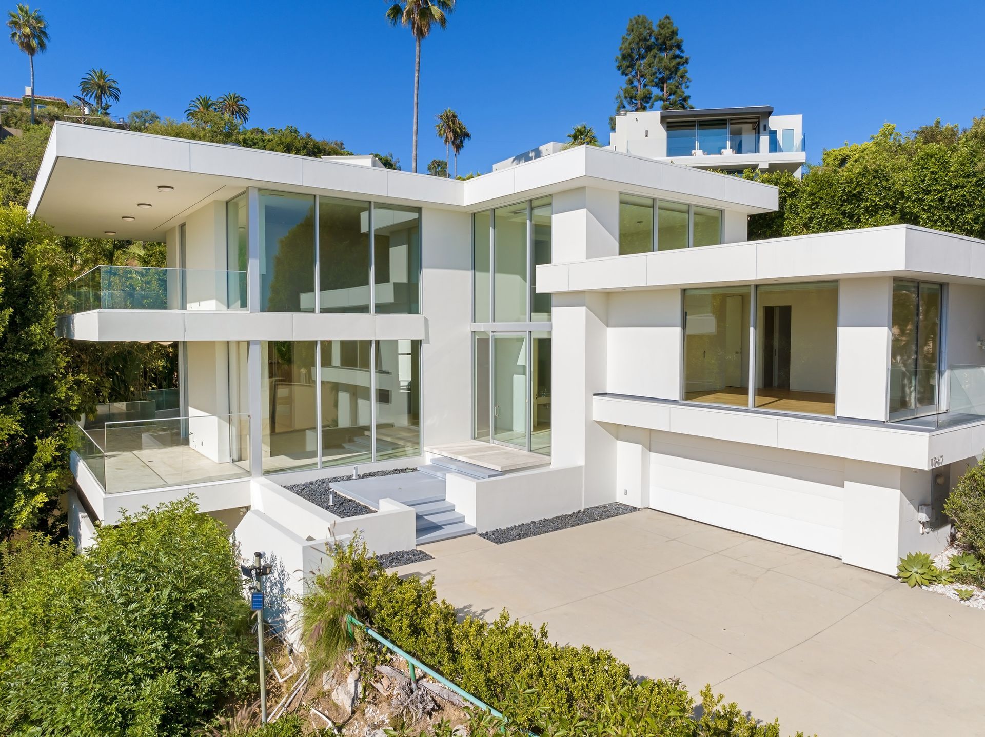 Modern white house with glass walls and balconies on a hillside, sunny day.