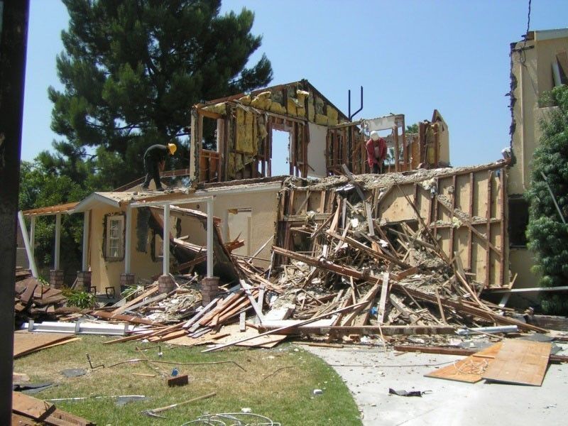 House under demolition; debris scattered in yard, workers on roof removing boards.