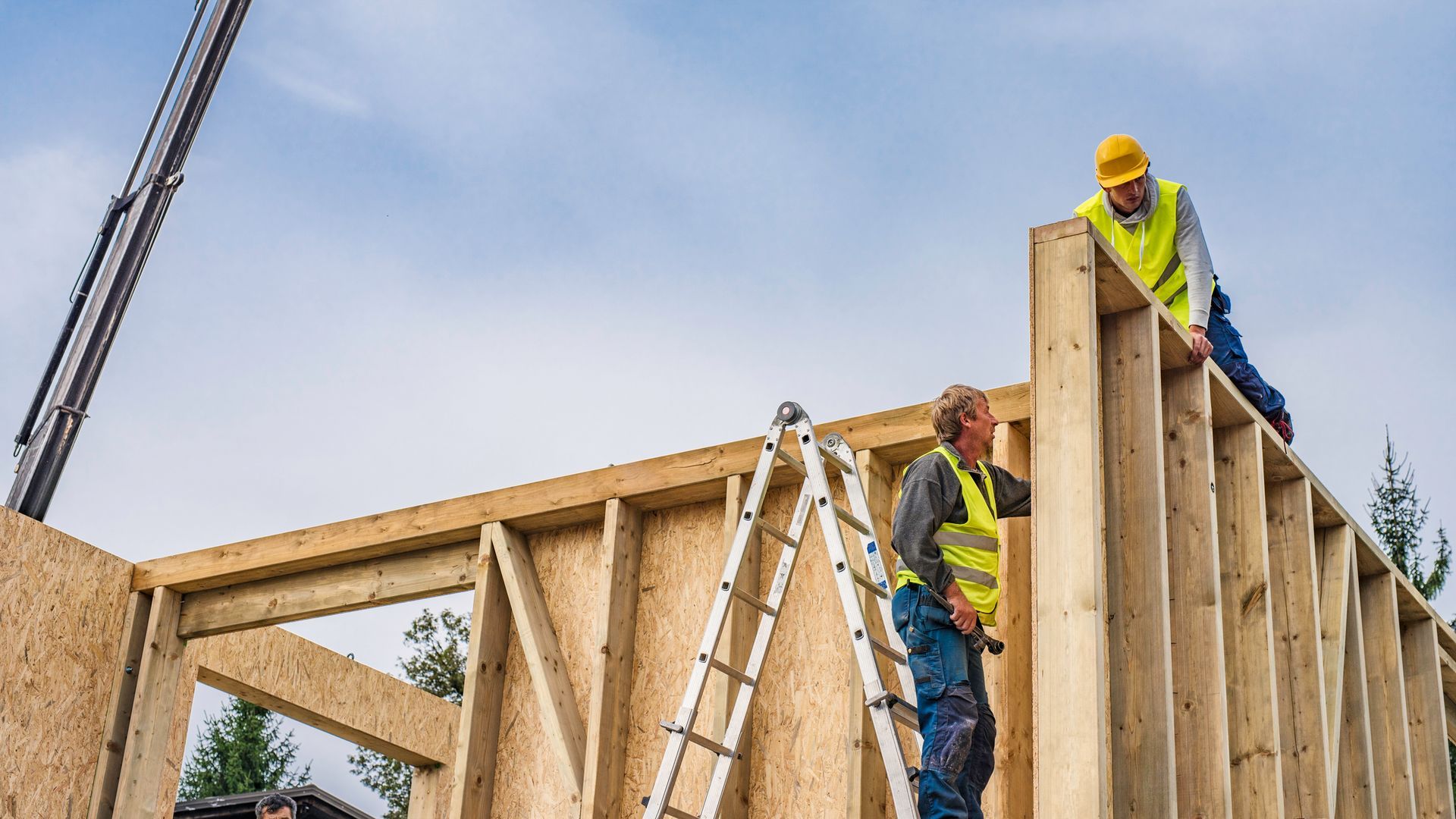 Construction workers building a wooden wall outdoors, wearing safety vests and helmets, under a partly cloudy sky.