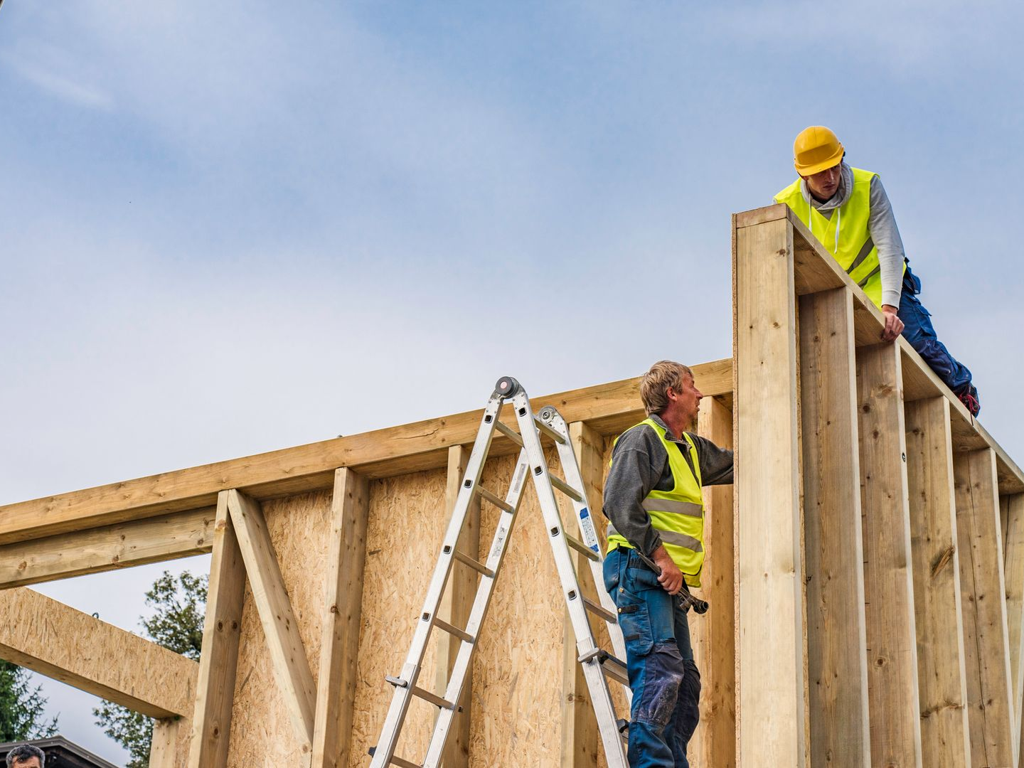 Construction workers building a wooden wall, wearing safety vests and hard hats, outdoors.