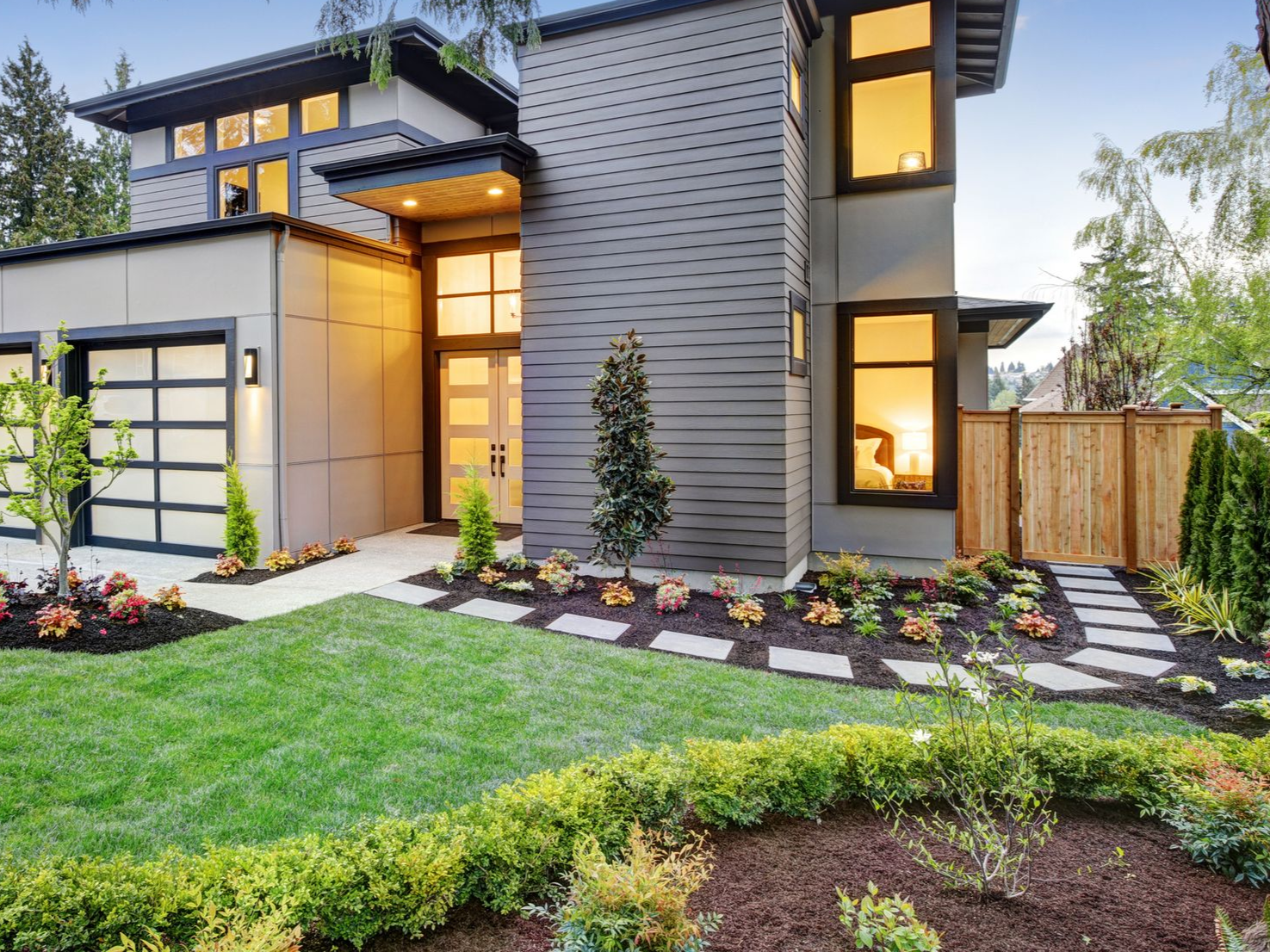 Modern two-story house with gray and tan siding, lush green lawn, and landscaped flower beds.