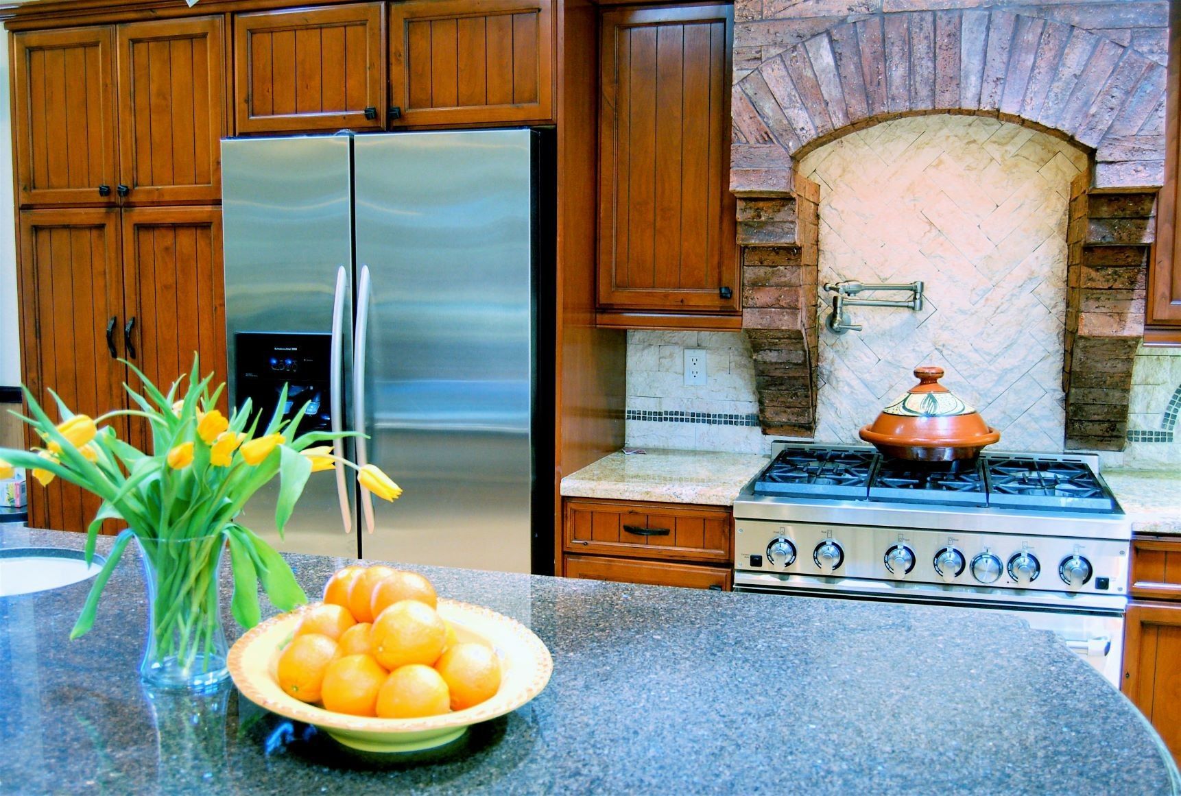 Kitchen with stainless steel refrigerator, wooden cabinets, and a brick-arched stove area. Fresh fruit and flowers on countertop.