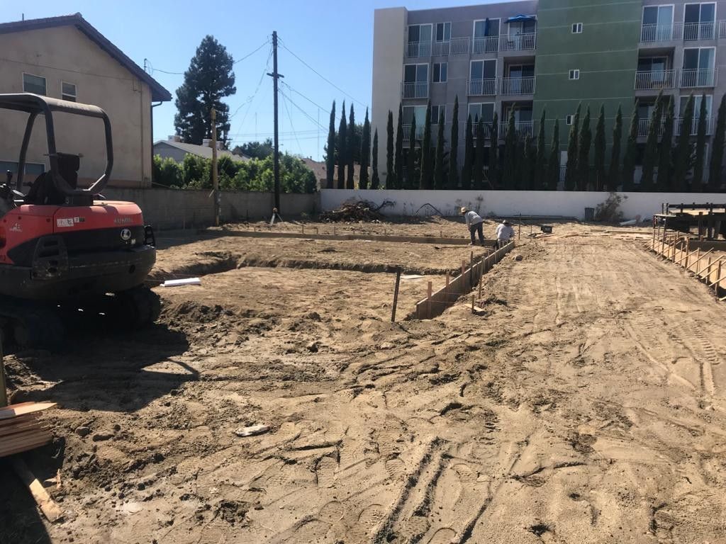 Construction site with mini-excavator, dirt, and concrete forms. A worker stands nearby a building under construction.