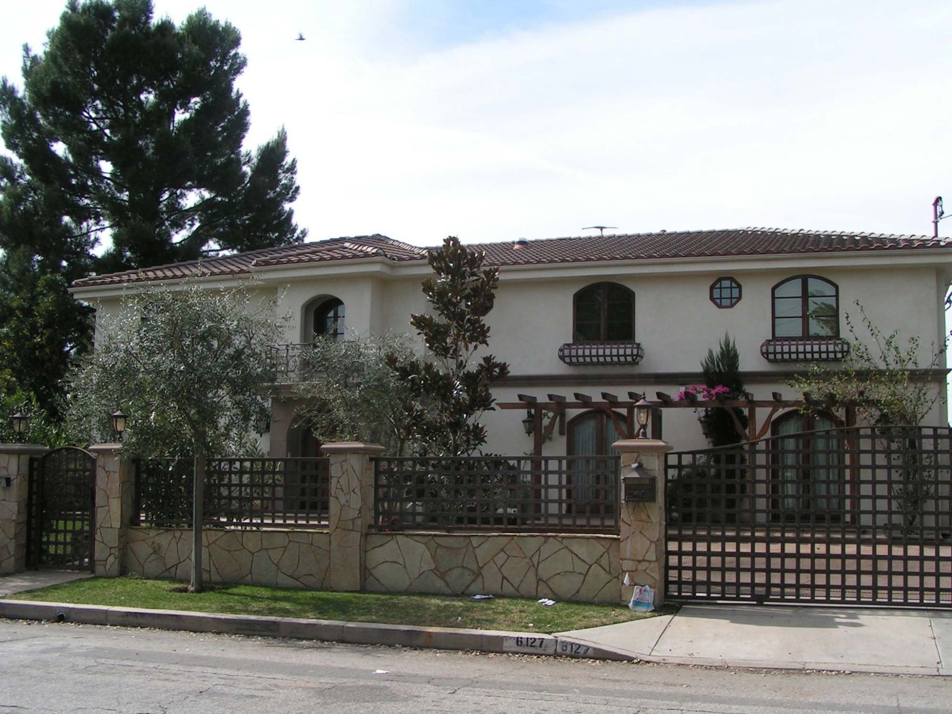 Two-story beige house with a gated entry, tiled roof, and arched windows. Trees and blue sky in the background.