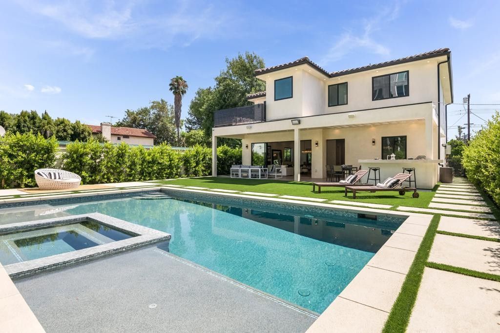 A two-story house with a pool and jacuzzi in the backyard. Someone is lounging on a chair.