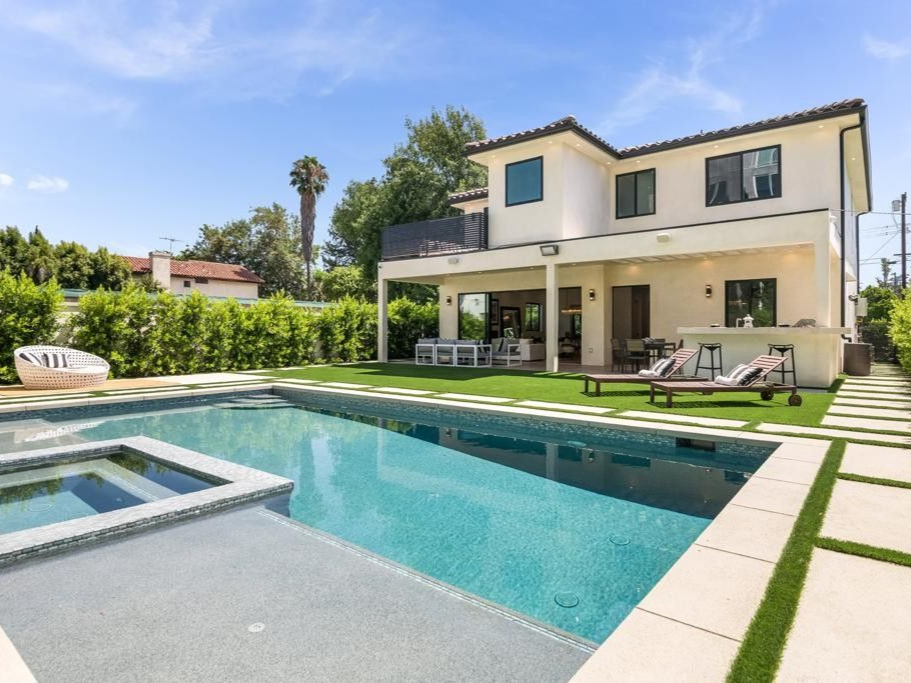 Two-story house with a pool and hot tub in a sunny backyard with grass and stone pavers.