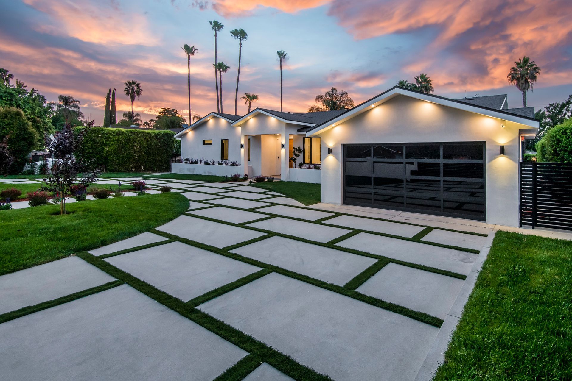 Modern white house with a landscaped yard and patterned driveway against a sunset sky.