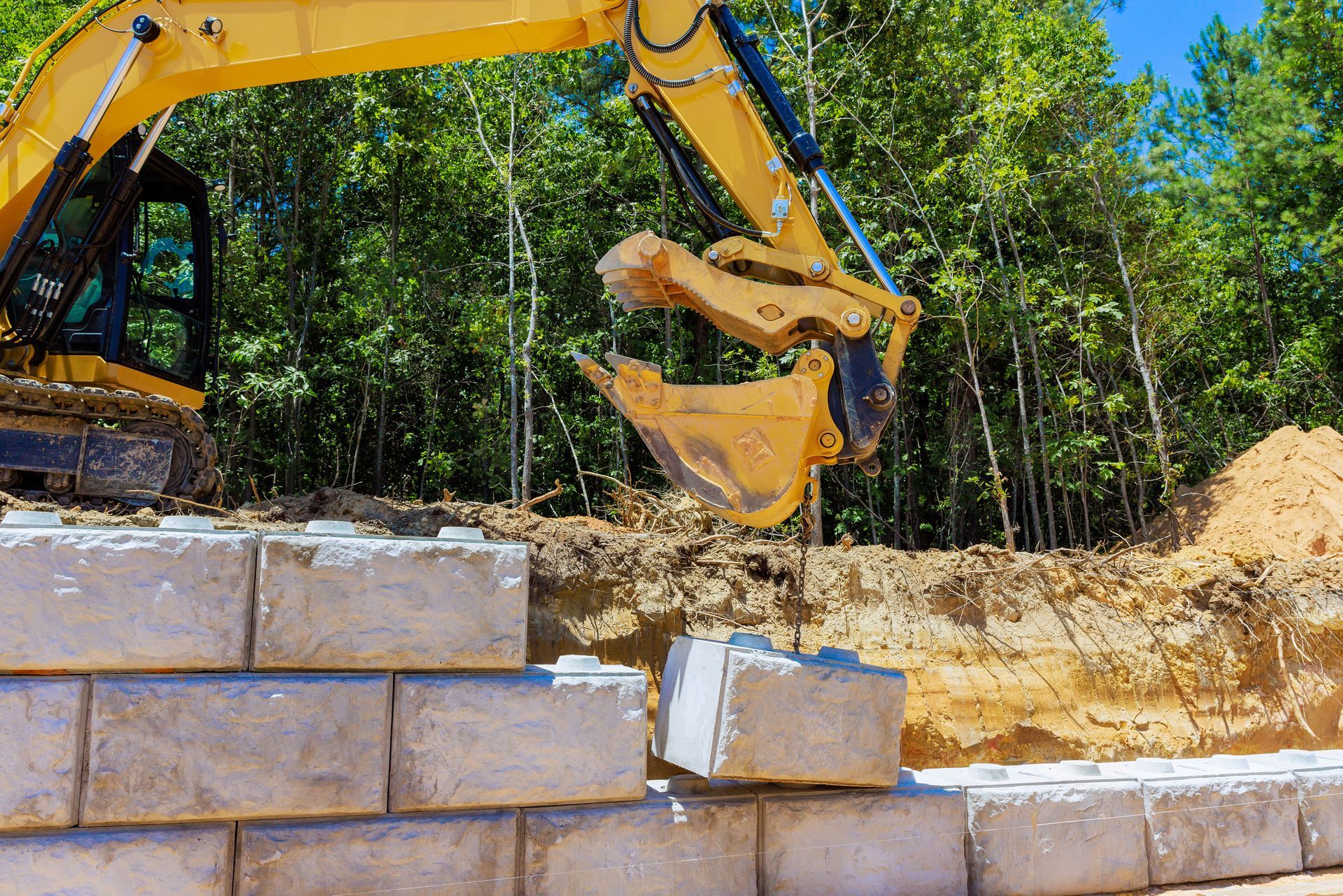 Yellow excavator placing a concrete block on a retaining wall under construction.