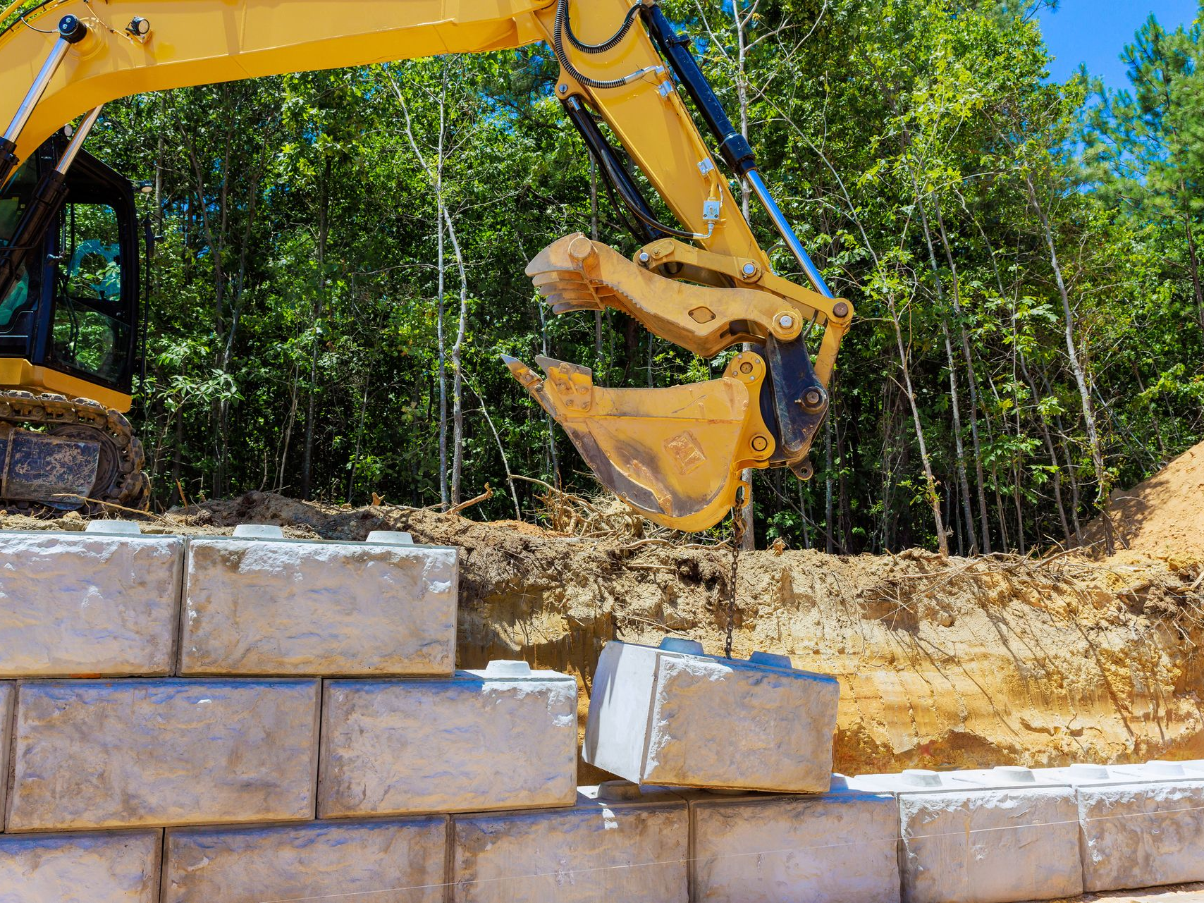 Excavator placing concrete block for retaining wall in construction zone near trees.