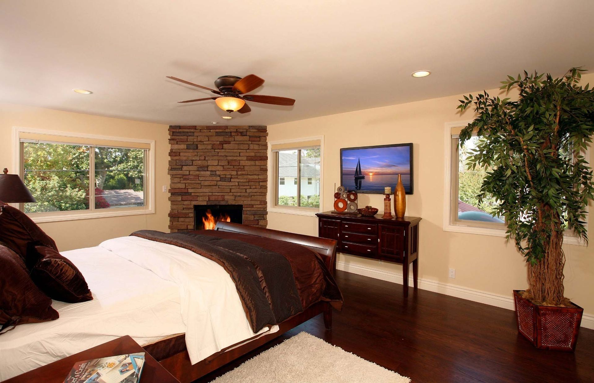 Spacious bedroom with fireplace, TV, and dark wood furniture. A large bed sits in the foreground with windows.