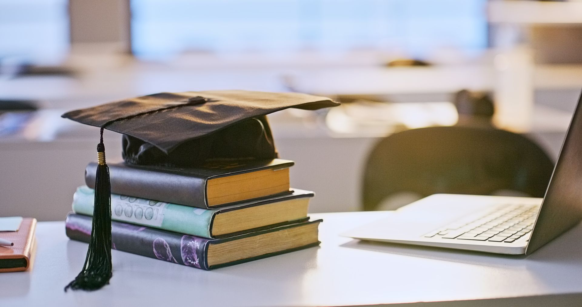 Graduation cap on a stack of books next to a laptop on a white desk.