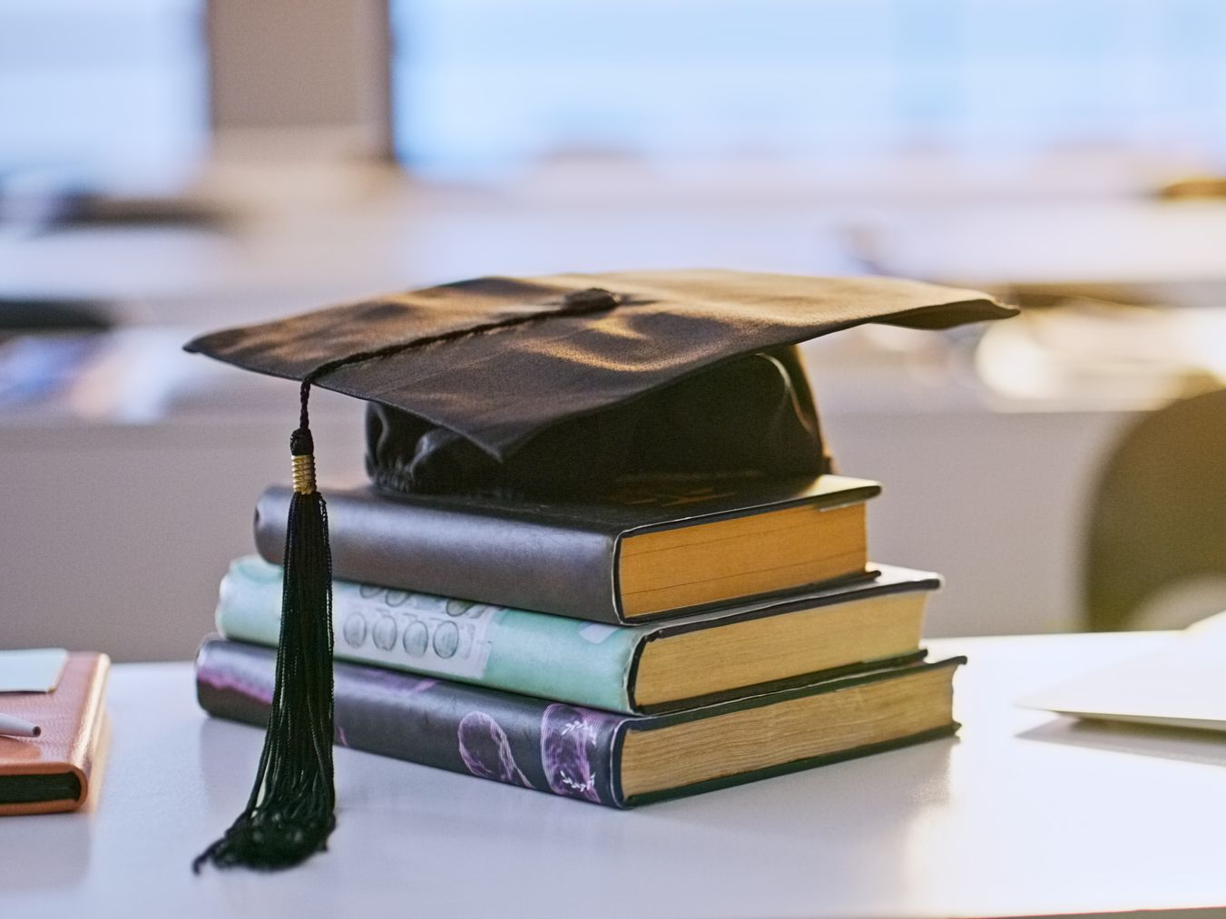 Graduation cap atop a stack of books on a desk; academic setting, soft lighting.