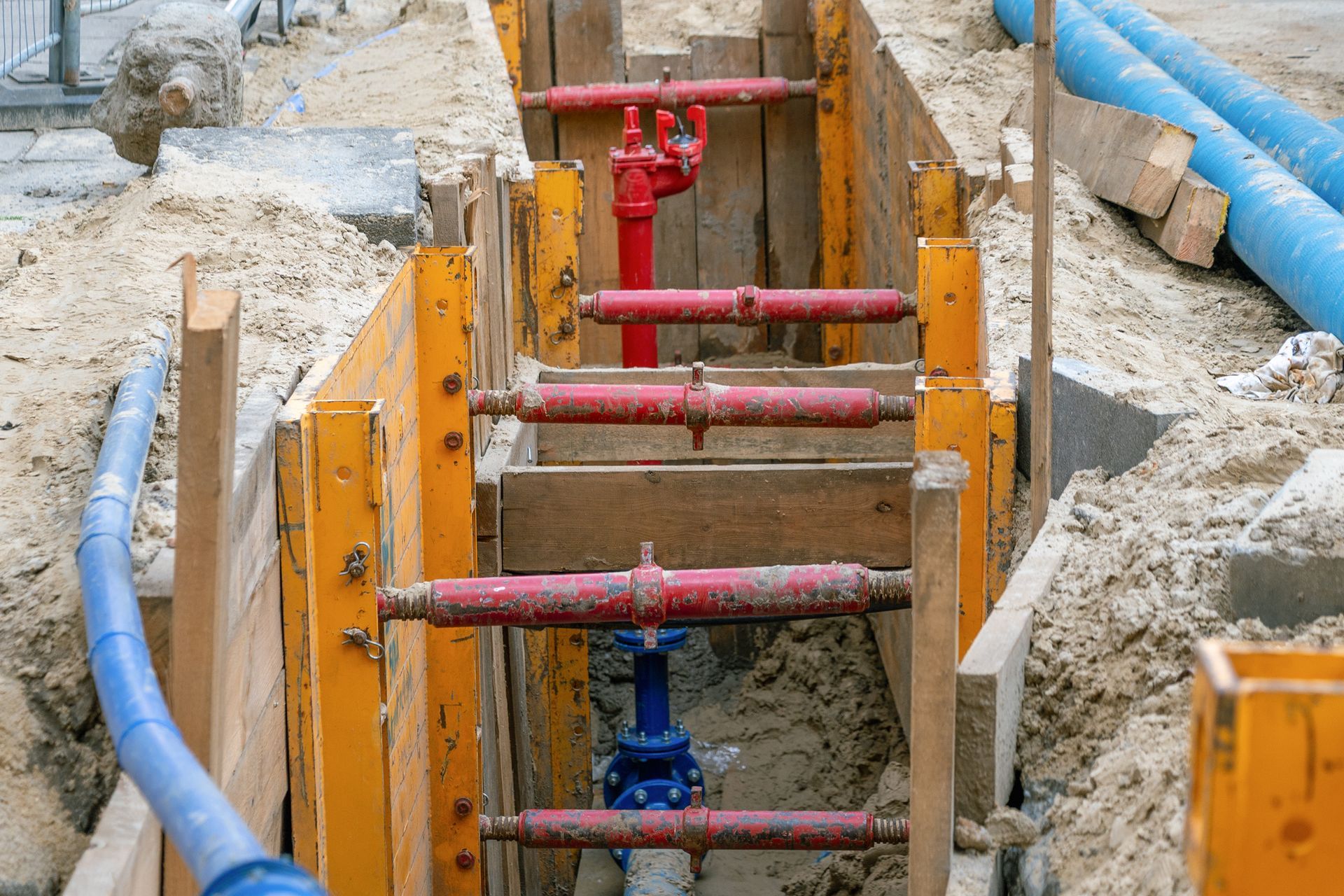 Trench with yellow shoring, red and blue pipes and valves, sandy soil, and blue corrugated pipe.