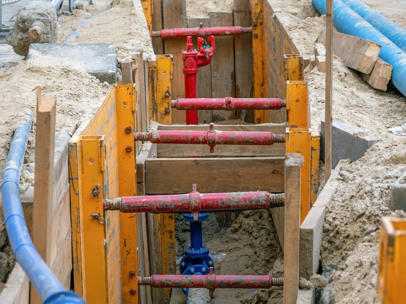 Trench with yellow shoring, red and blue pipes and valves, sand, and blue pipe partially visible.