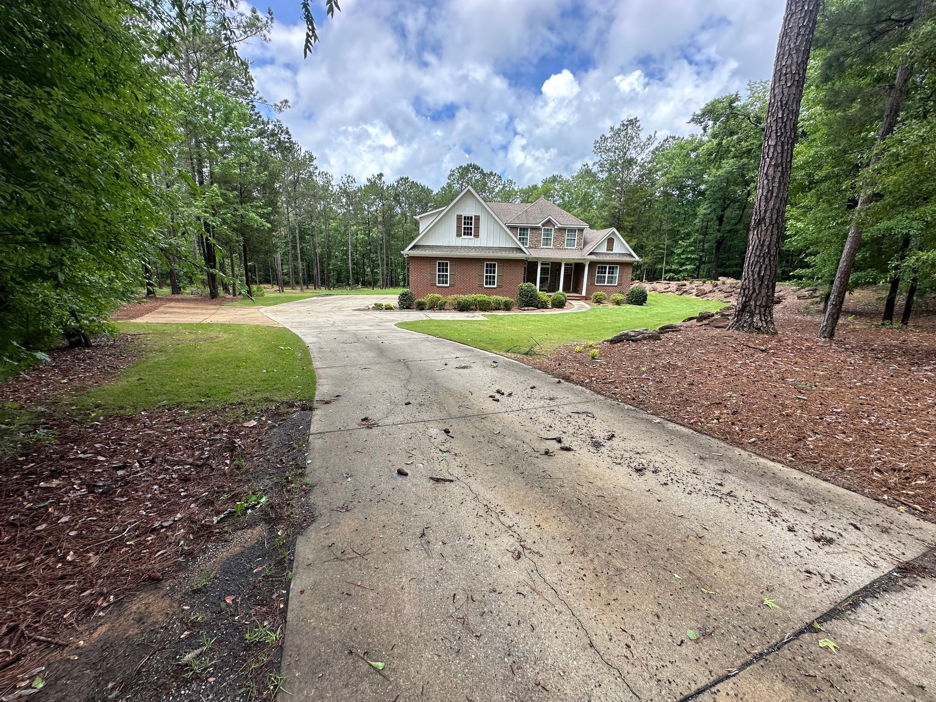 Brick house with a long driveway surrounded by trees and greenery under a cloudy sky