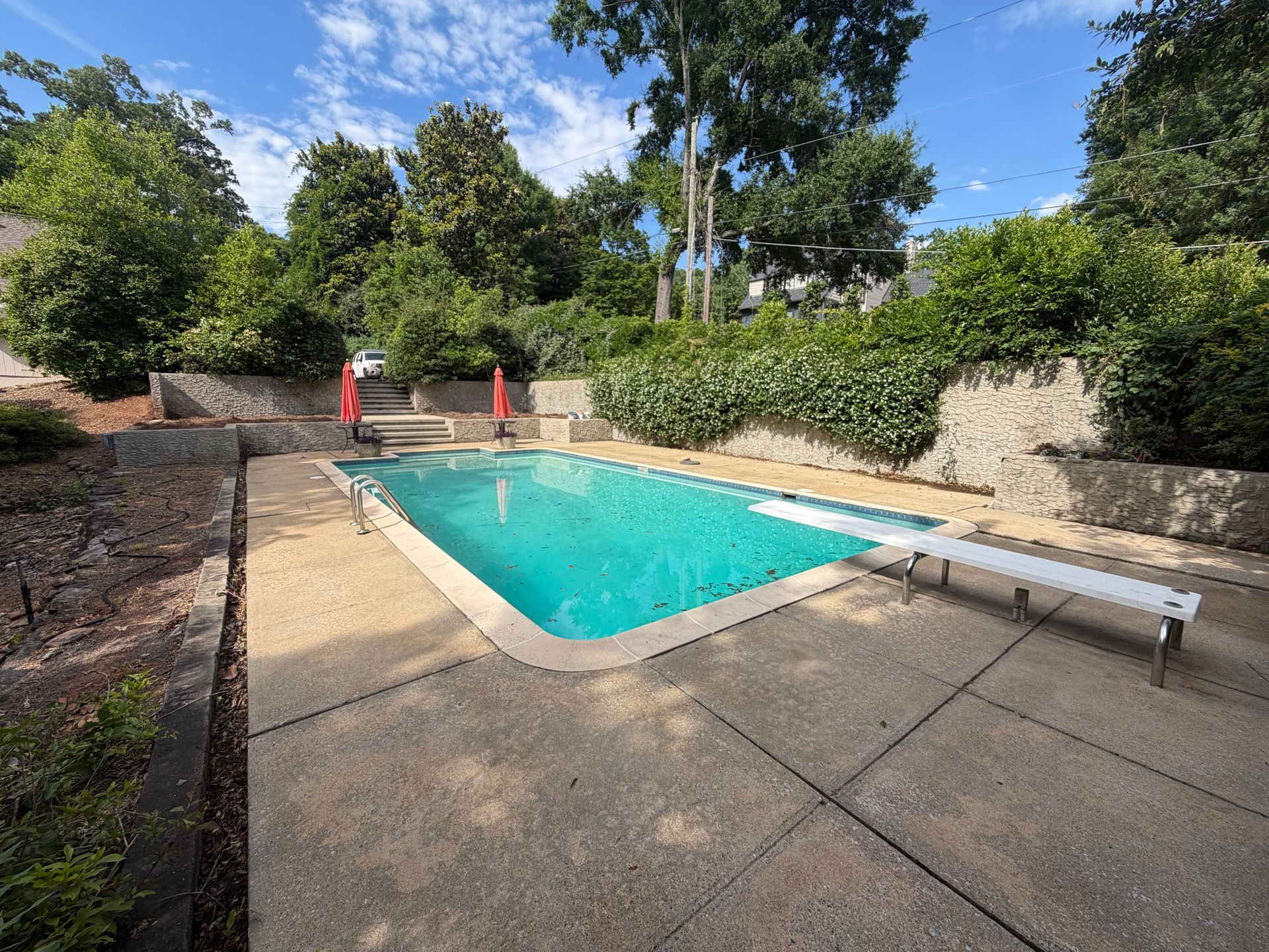 Backyard pool with turquoise water, concrete deck, lounge chair, and red umbrellas surrounded by trees and shrubs