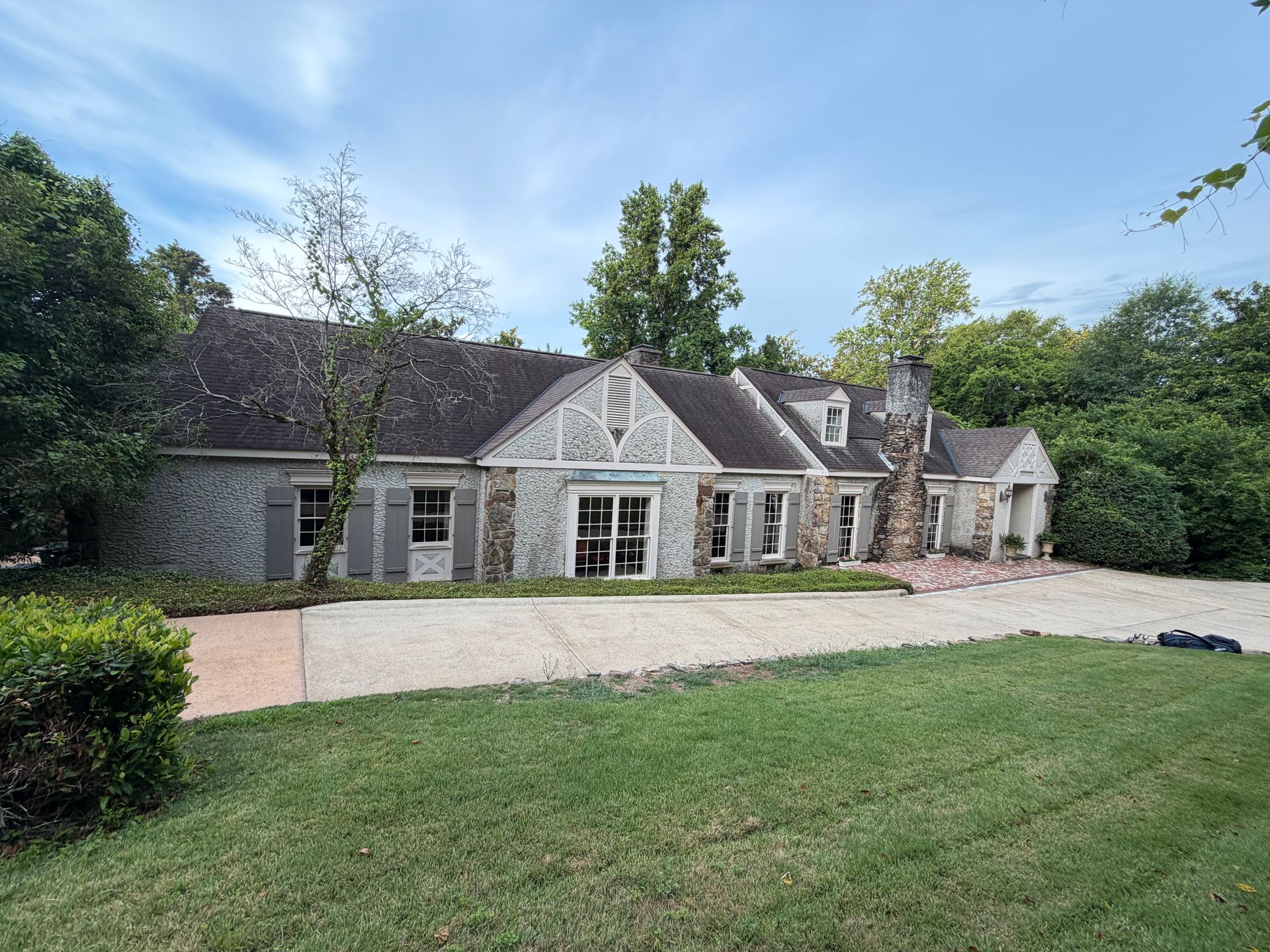 Stone cottage with a gravel driveway and green lawn, surrounded by trees under a cloudy sky