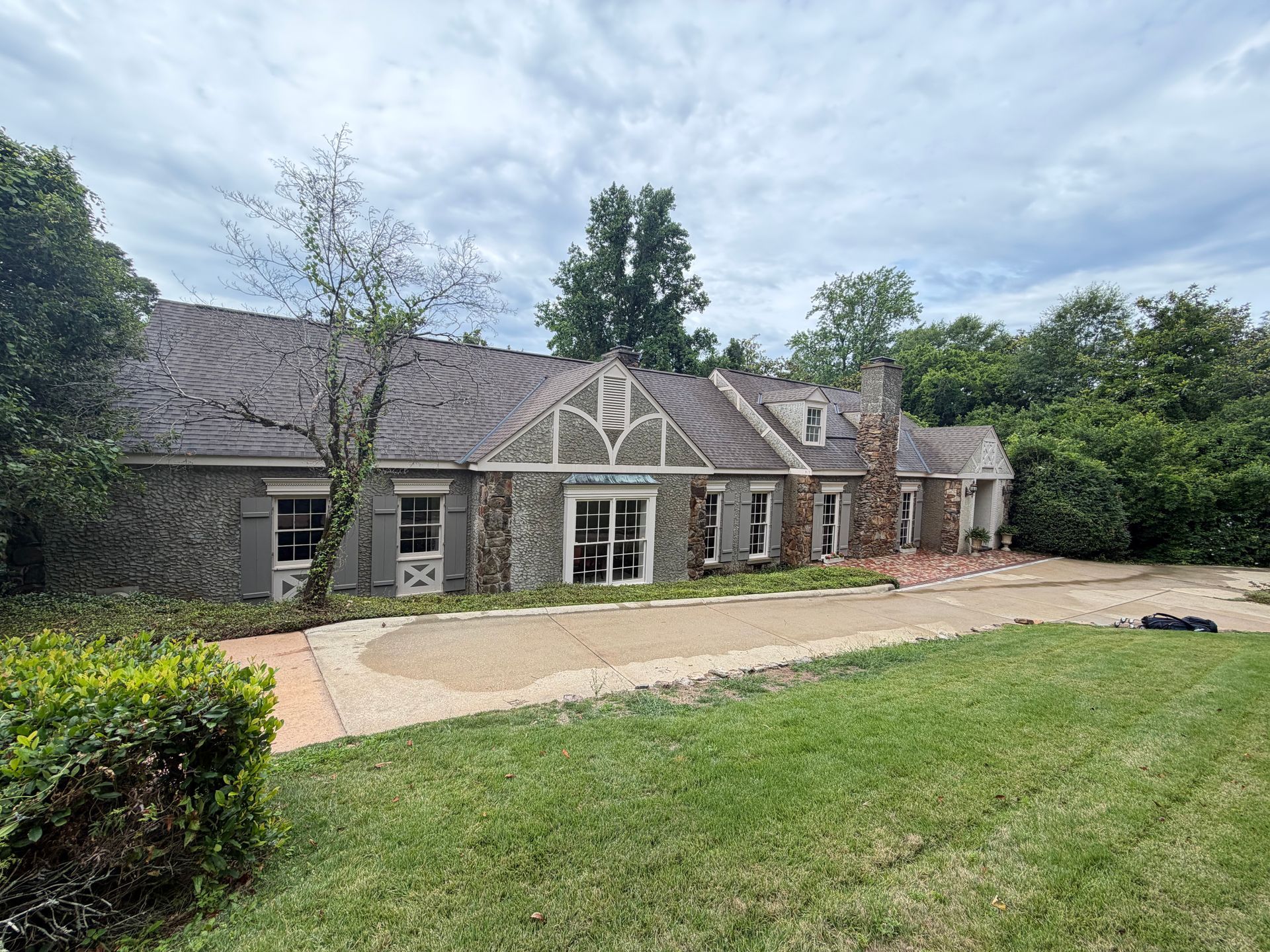 Stone suburban house with gray roof, large windows, and a wide lawn under a cloudy sky
