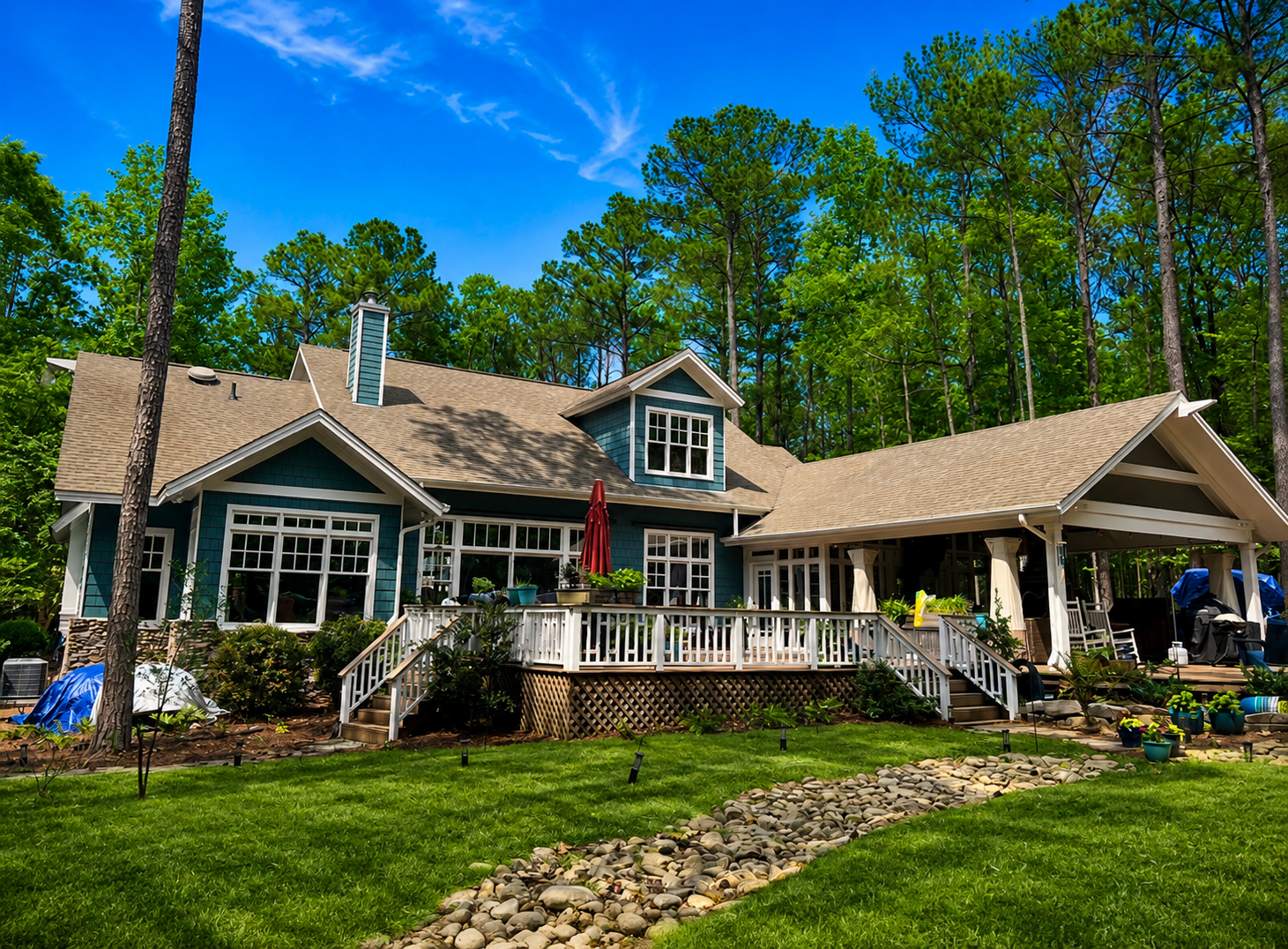A gray cottage with white trim and a porch, surrounded by green lawn and tall trees under a bright blue sky