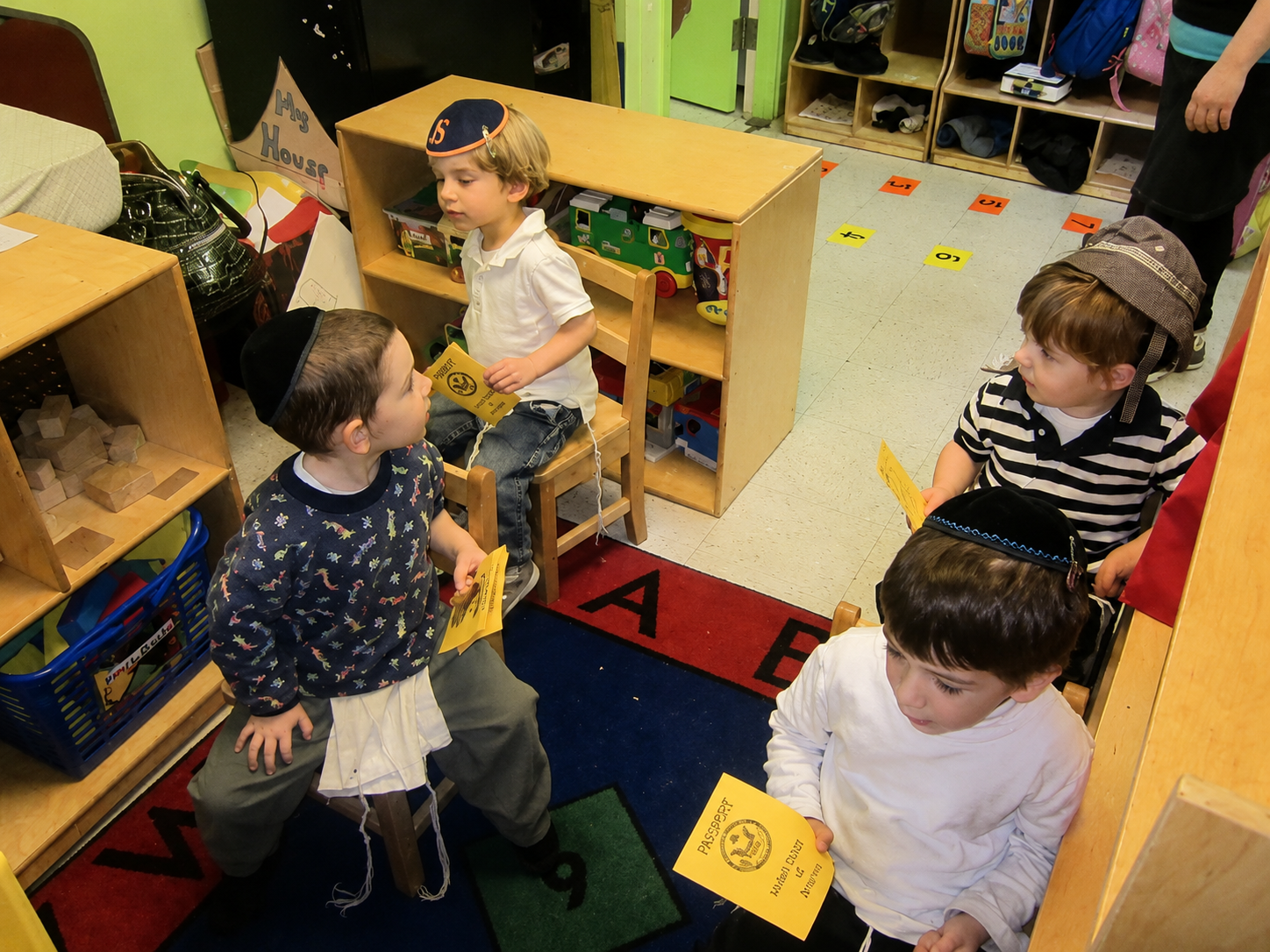 Children sitting on the floor in a colorful classroom, holding papers and talking near low shelves.