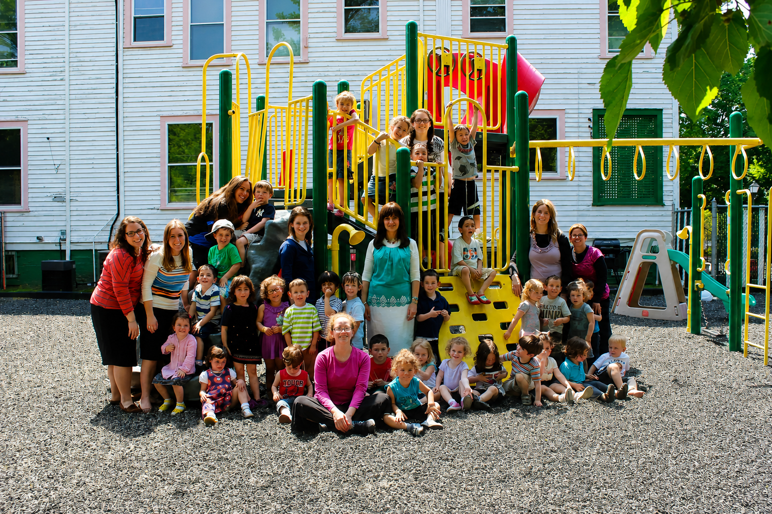 Large group gathered at a colorful playground, posing on gravel in front of play structures and a building.