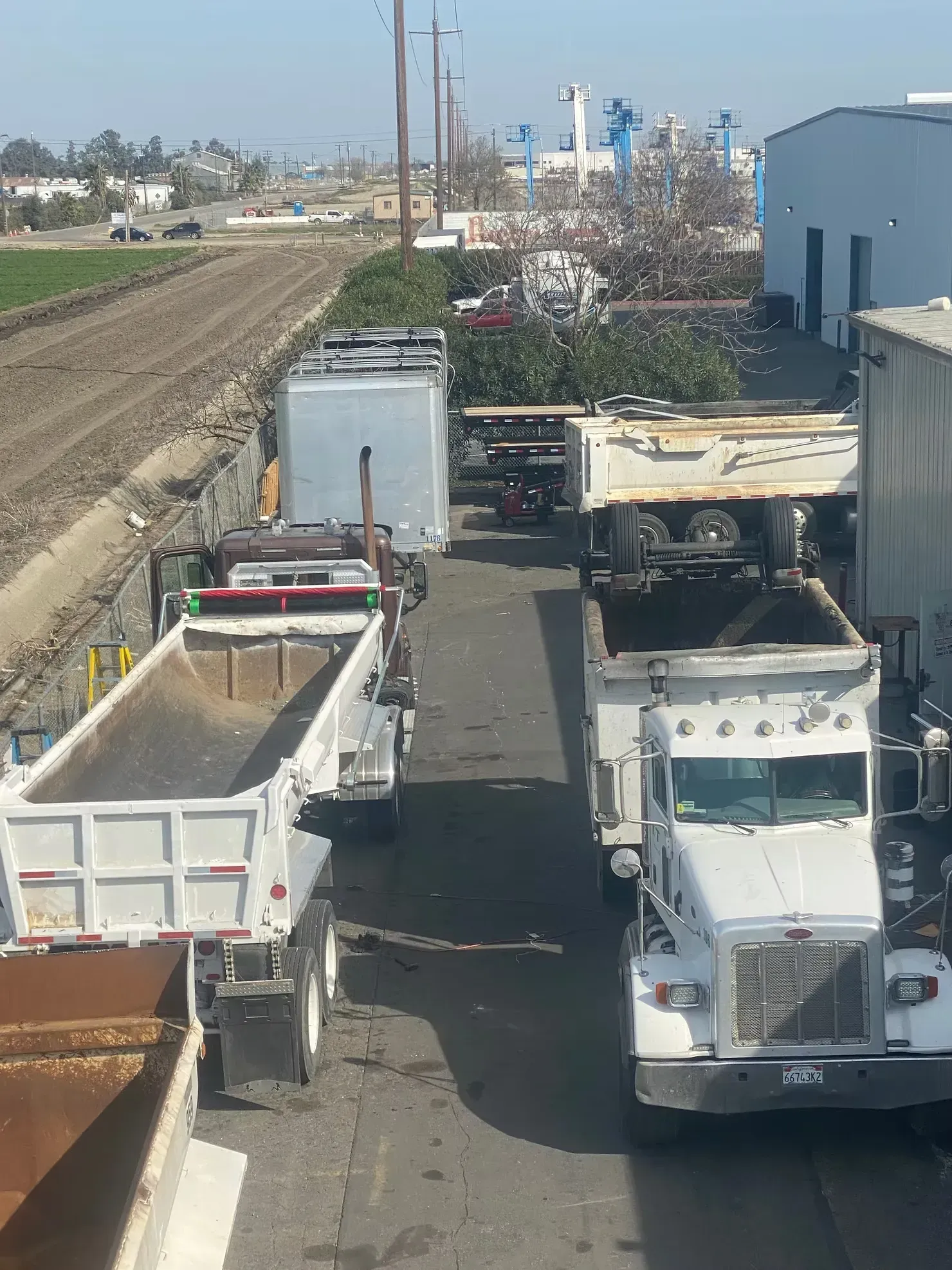 Dump trucks lined up on a roadside beside a dirt field and construction area.
