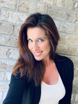 A woman with long brown hair is smiling in front of a brick wall.