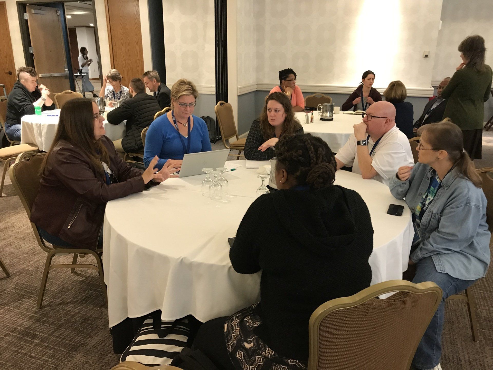 People seated at round tables in a conference room, engaged in discussion. Some have laptops.