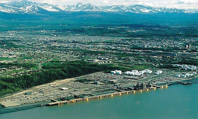 Aerial view of a coastal city with a port, mountains in the background, and blue water in the foreground.