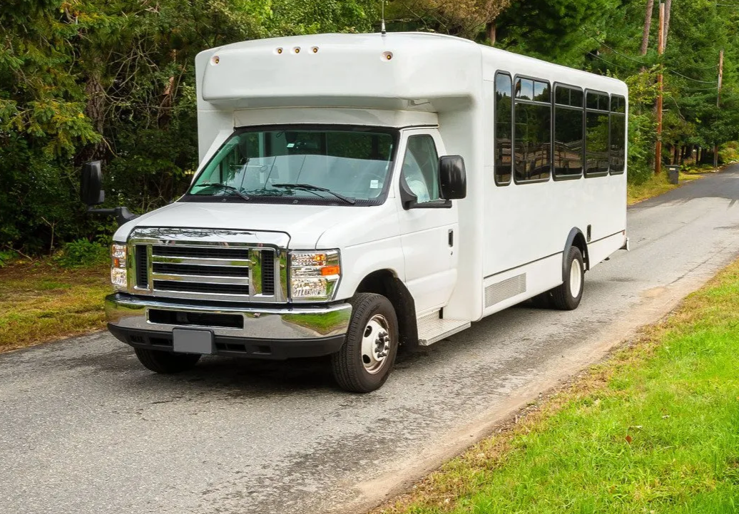 White shuttle bus parked on a gravel road beside green trees and grass