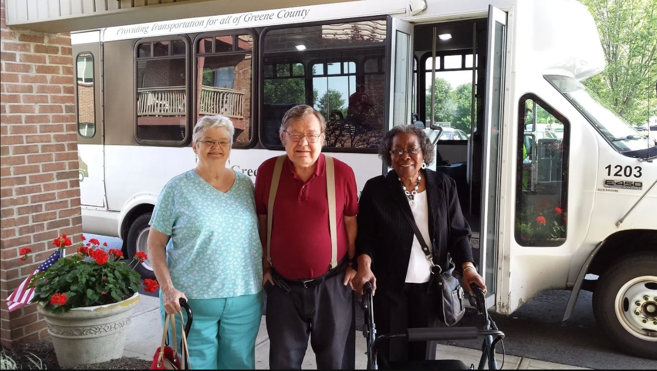 Three older adults pose next to a white transport van. One uses a walker, flowers in foreground.