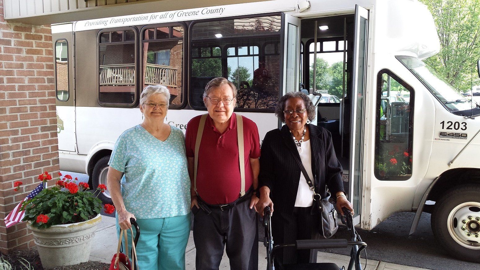 Photo of 3 people standing in front a a bus.  one is using a walker.