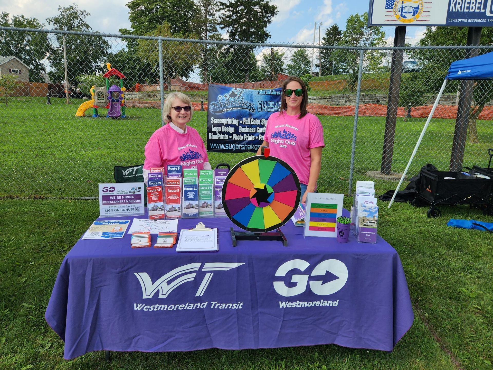 Two women are standing behind a purple table with a rainbow wheel and brochures on it.