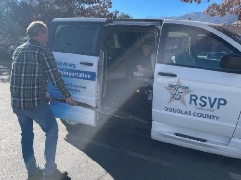 Group of seniors standing next to a transport van. Sunny day. Some use canes. Phone number on van.