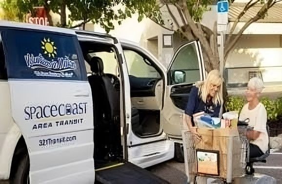 White van with a ramp, two women loading groceries. Space Coast Area Transit van, outdoor setting.