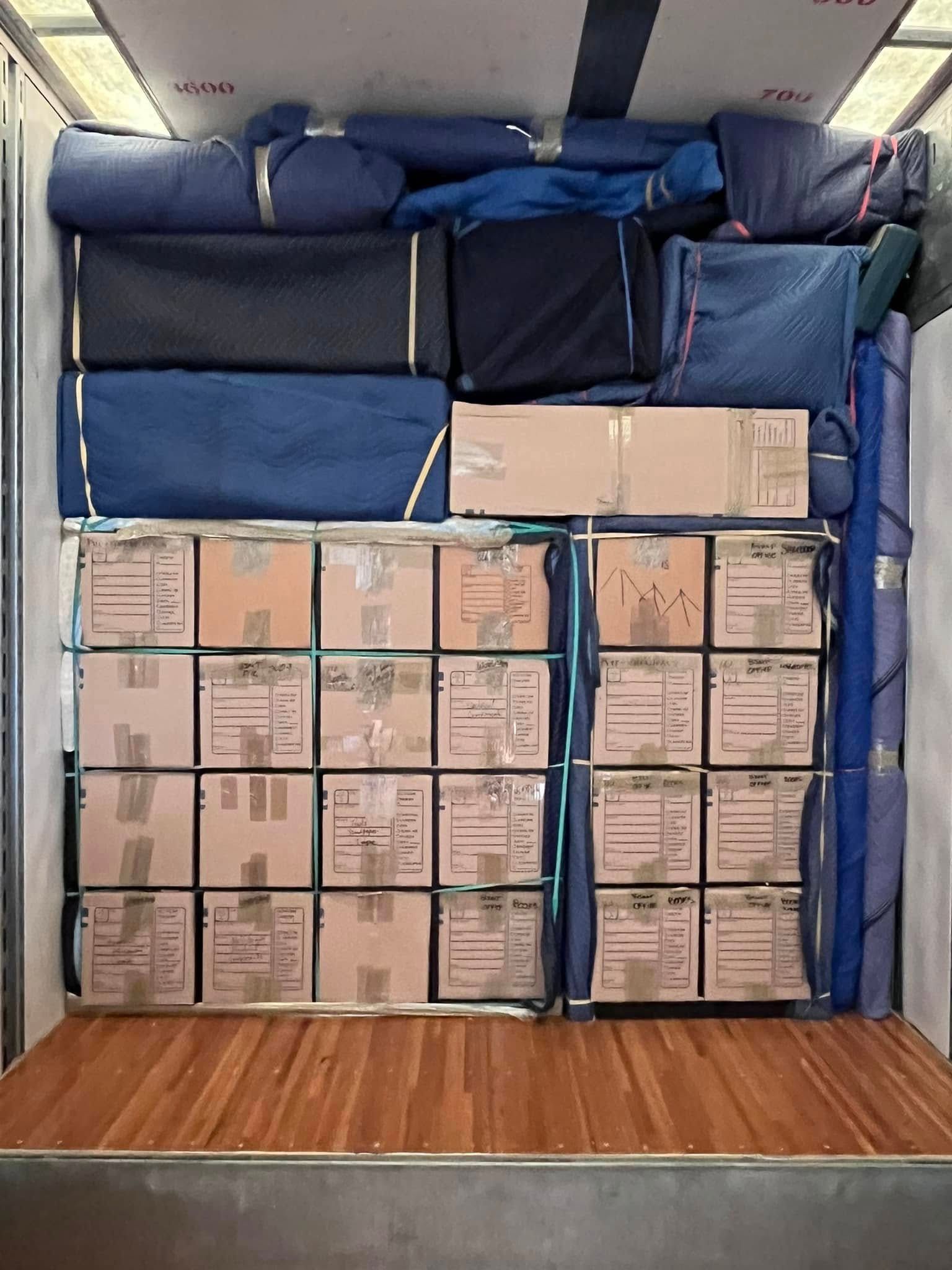 Two movers in blue uniforms and gloves carry a grey sofa across a bright living room with wood floors.