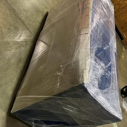 Three workers in blue shirts and tan pants move a sofa and wooden table from a moving truck on a city street.