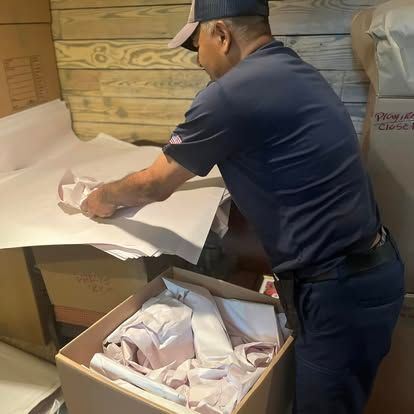 Two workers in blue uniforms unloading brown cardboard boxes from the back of a white delivery van parked outside.