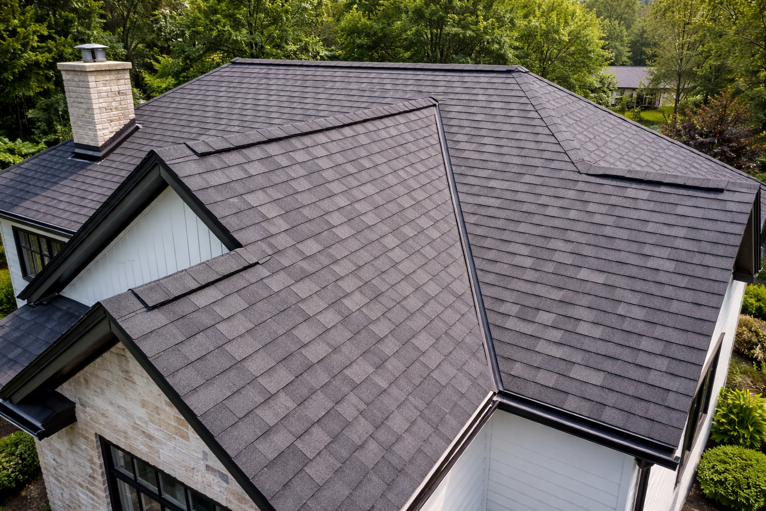 Dark gray shingle roof on a house, angled view, chimney, greenery in background.