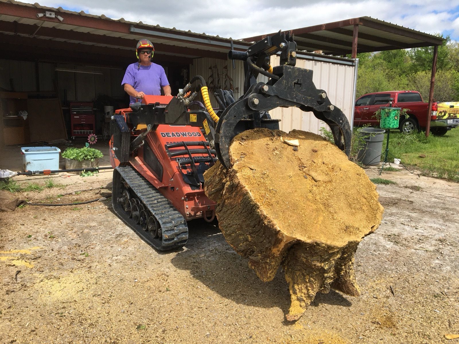 A person operating an orange skid steer with a grapple lifting a large tree stump in an outdoor setting.