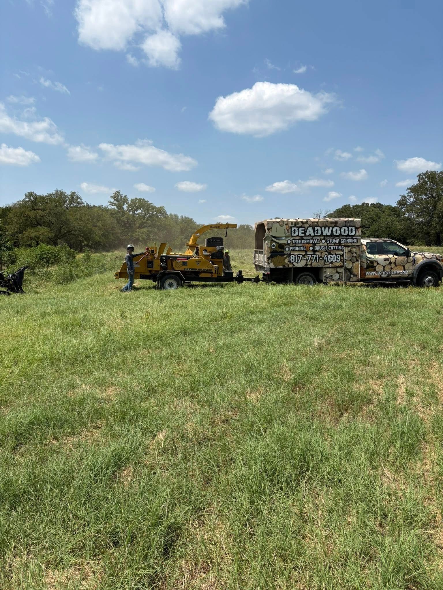 A wood chipper and work truck in a grassy field under a blue sky, with trees in the background.
