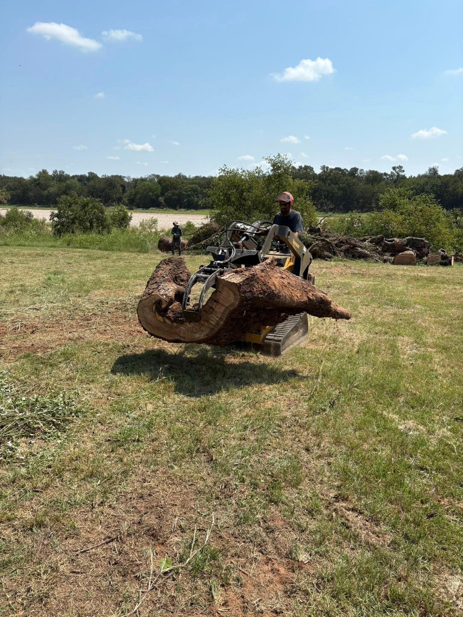 Person operating a small tractor carrying a large, textured piece of wood in a grassy field.